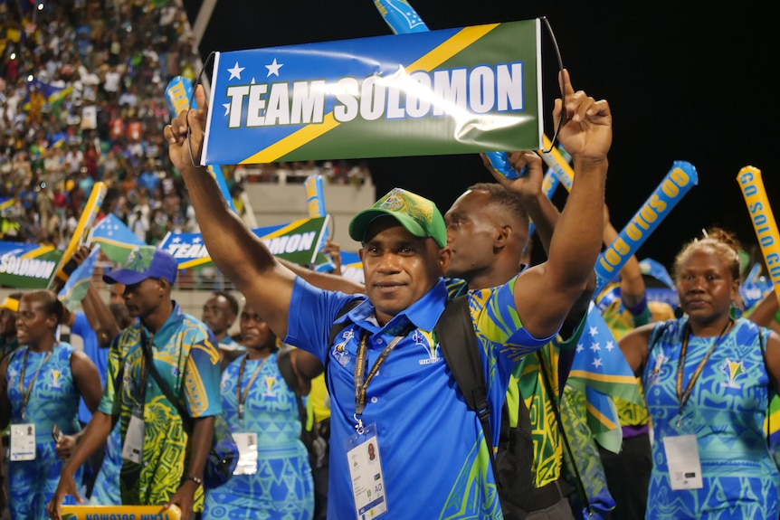 A man proudly holds a sign reading Team Solomon, as he and other wear blue, yellow and green colored uniforms.