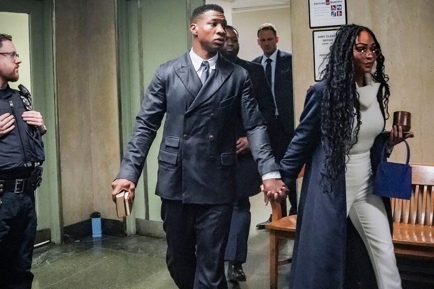 Jonathan Majors in leather suit enters courtroom holding hands with girlfriend Megan Good. 