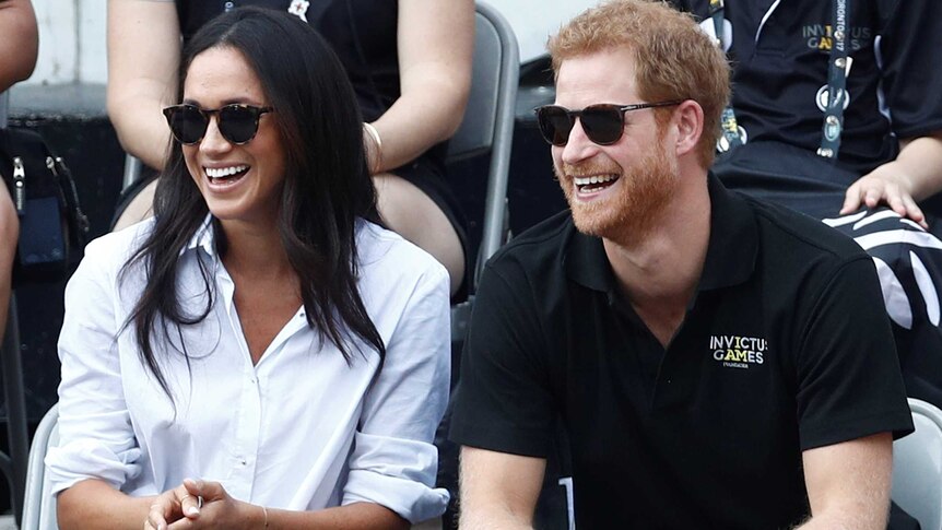 Meghan Markle wearing a white shirt and jeans and Prince Harry in a black T-Shirt watching wheelchair tennis from the crowd.