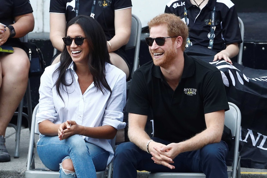 Meghan Markle wearing a white shirt and jeans and Prince Harry in a black T-Shirt watching wheelchair tennis from the crowd.