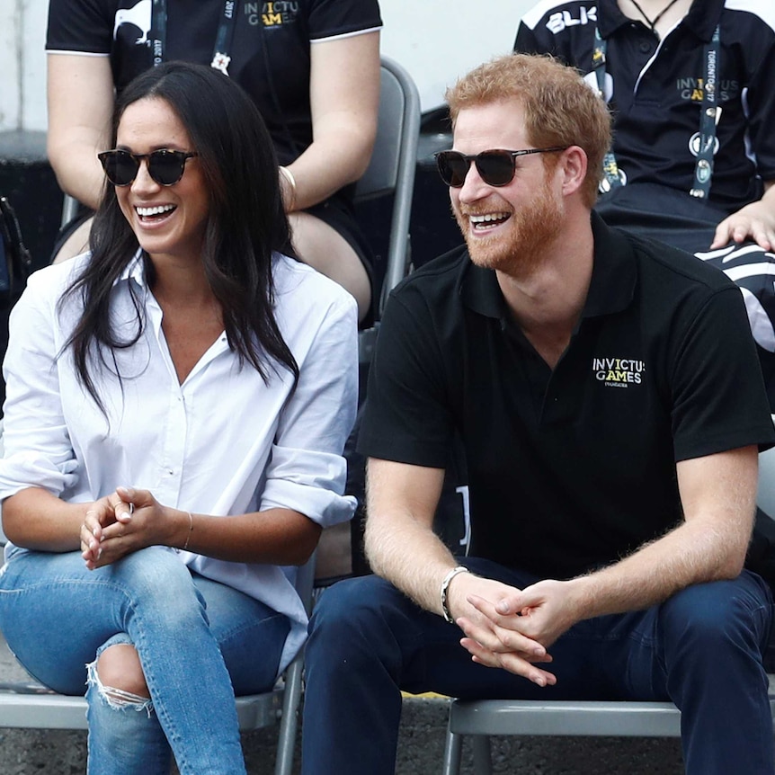 Meghan Markle wearing a white shirt and jeans and Prince Harry in a black T-Shirt watching wheelchair tennis from the crowd.