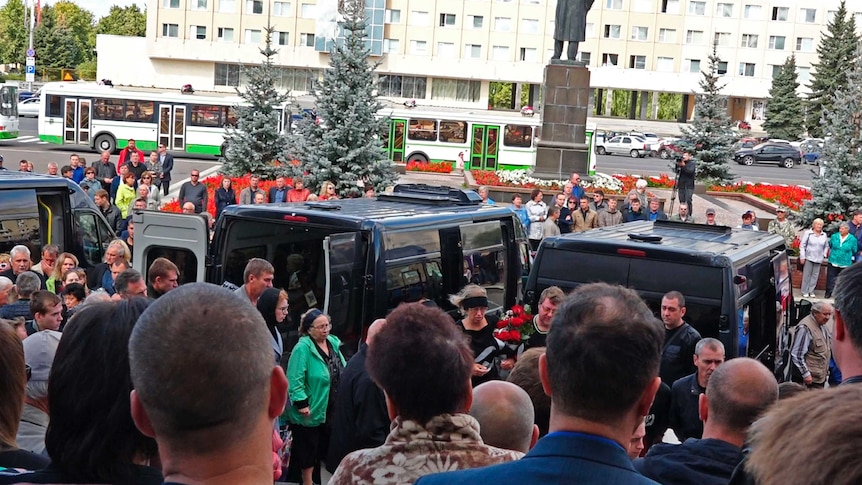 A large number of people, walking in front of black vans, go to the funeral service