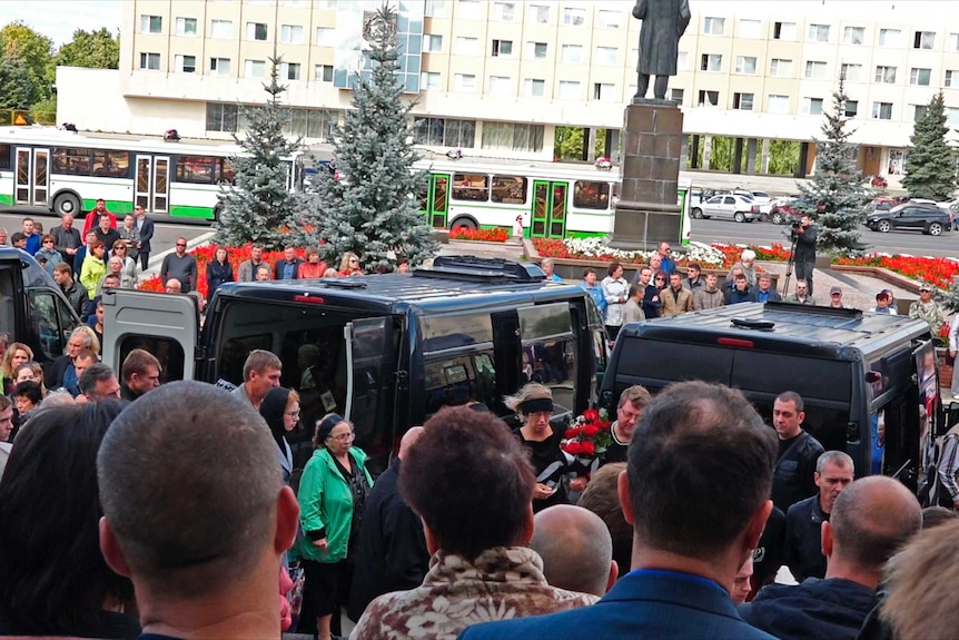 People gather around five funeral cars in a street.