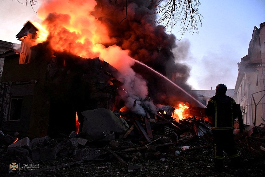 The silhouette of a firefighter stands before a large orange fire rising above rubble in evening light