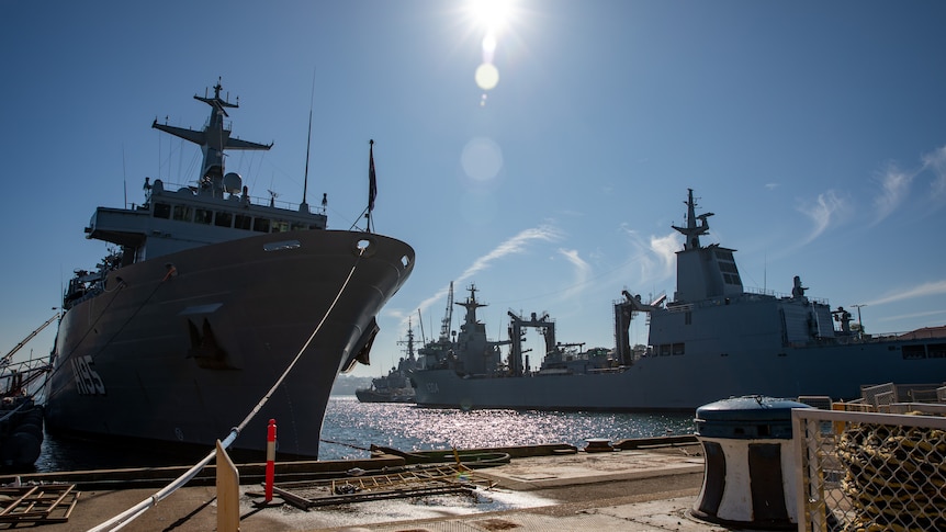 Two large military ships are docked with the sun in the background