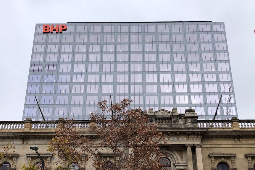 Un gran edificio rectangular moderno con el logotipo de BHP detrás de un edificio de piedra del siglo XIX.
