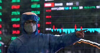 A man in a blue hazmat suit with a facemask and goggles stands in front of a stock exchange board.