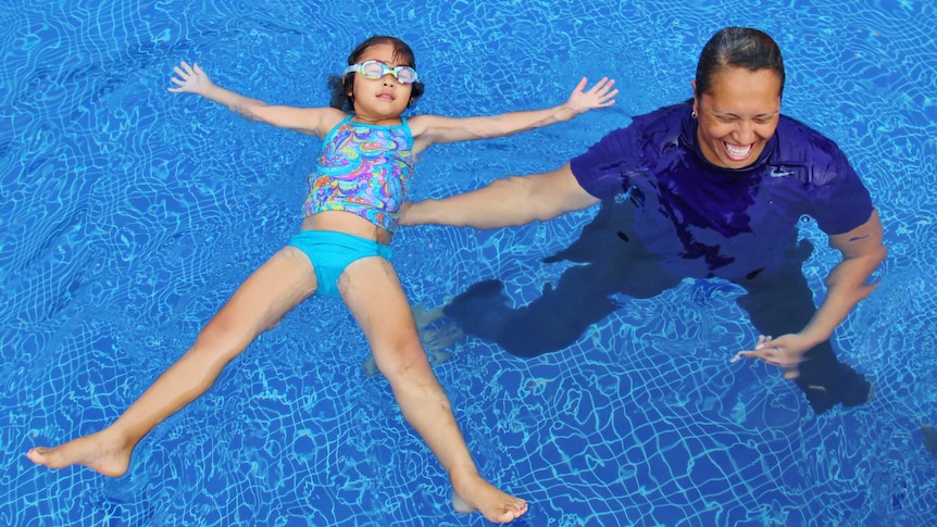 A young girl in blue swimmer floats on her back with arms and legs spread and her female coach is laughing.