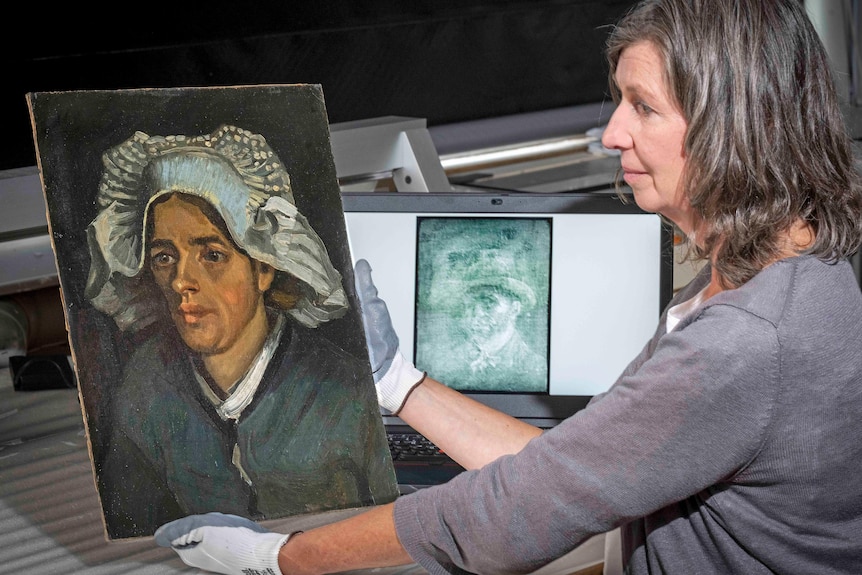 A woman wearing gloves holds a painting of a woman. A computer showing X-Ray of a man's painting in background.