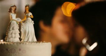 A cake topper of two brides while a same-sex couple kiss in the background.