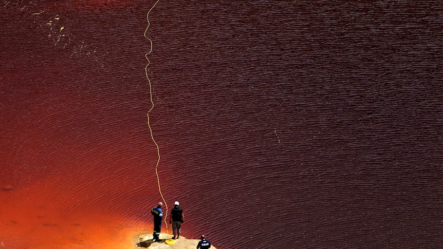 Three people stand on a rock as a blood-red lake ripples out in front of them. They stand by a long yellow cord flung into water