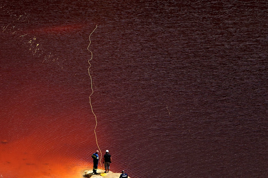 Three people stand on a rock as a blood-red lake ripples out in front of them. They stand by a long yellow cord flung into water