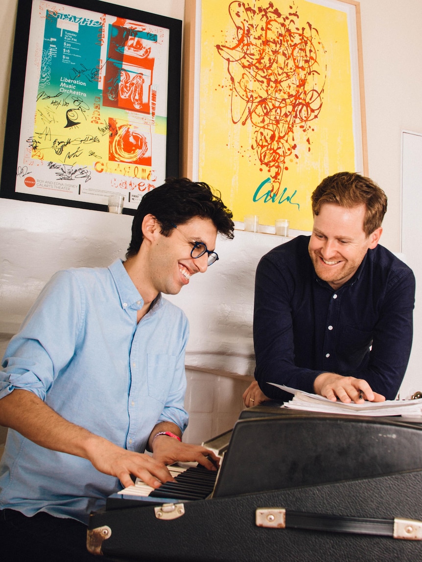Two men laughing. One is playing the piano, the other is leaning on it and a stack of papers