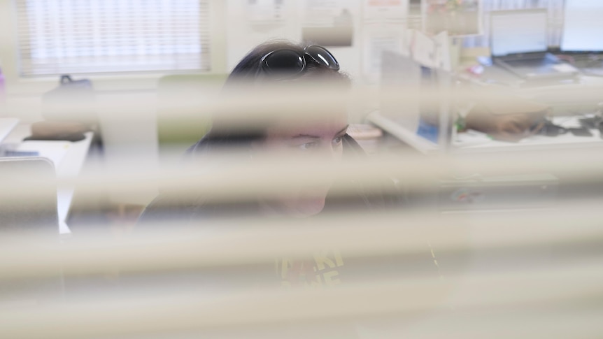 Lauren has black hair and is seen through the blinds of her office looking at her computer screen.