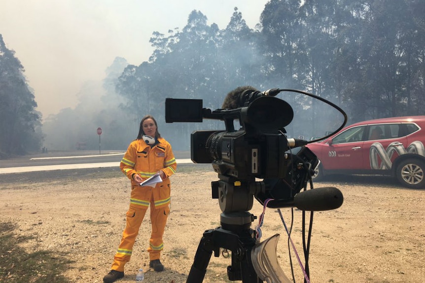 Reporter in yellow protective fire suit standing in front of camera next to ABC car with smoky haze in background.