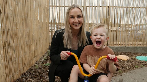 Young boy sitting on a tricycle is balanced by his mother. They are both smiling widely.