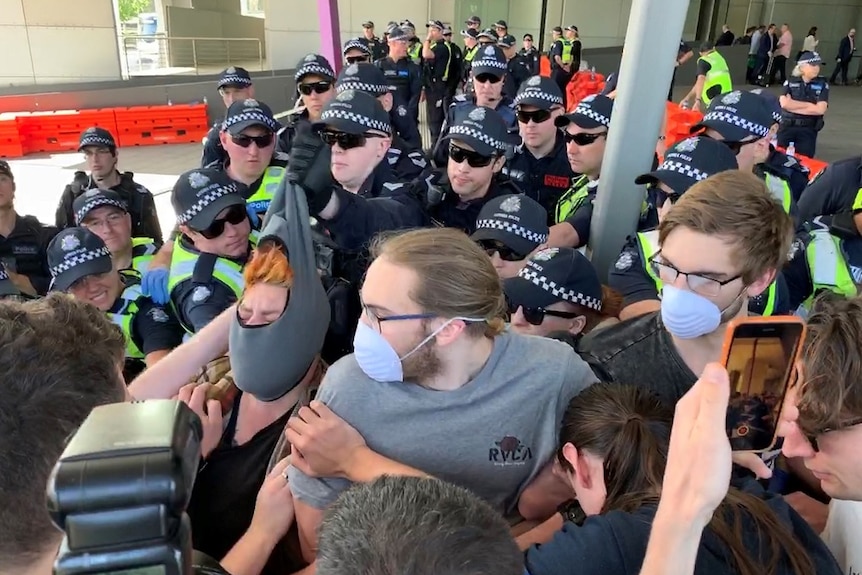 Police surround protesters who are wearing masks, one officer is seen pulling a balaclava off someone's face.