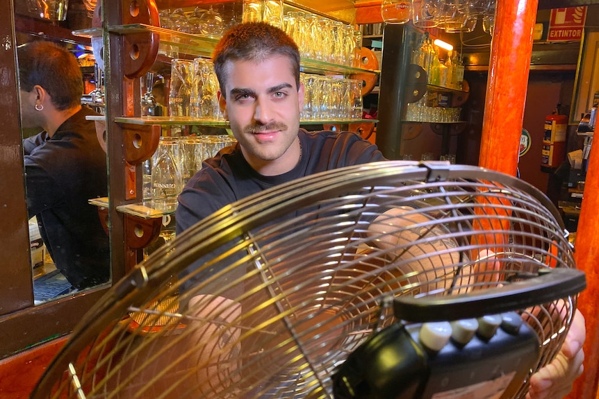 A barman with a moustache smiles as he cools down with a fan at a bar.