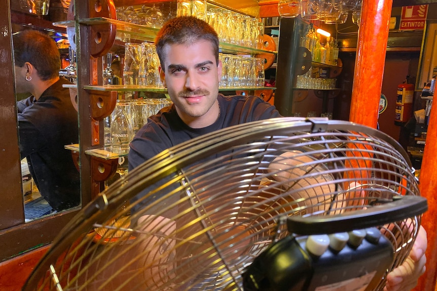 Camarero con bigote sonriendo mientras se refresca con un ventilador en el bar.