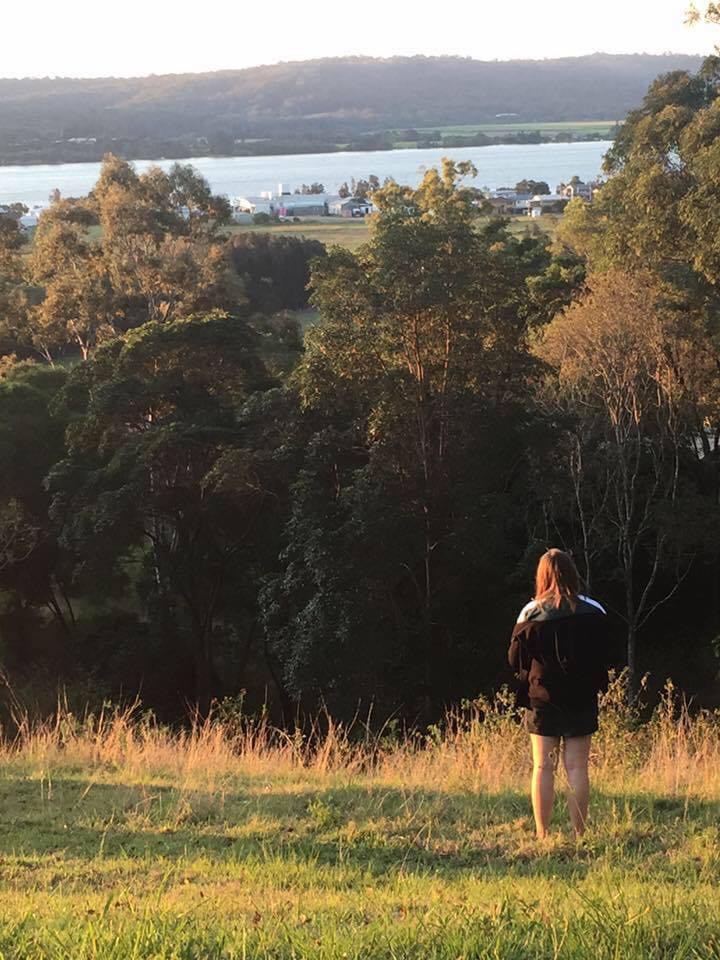 A woman, pictured from behind, stands in a grassy paddock. There is a distant view of a river and tree-covered hills.