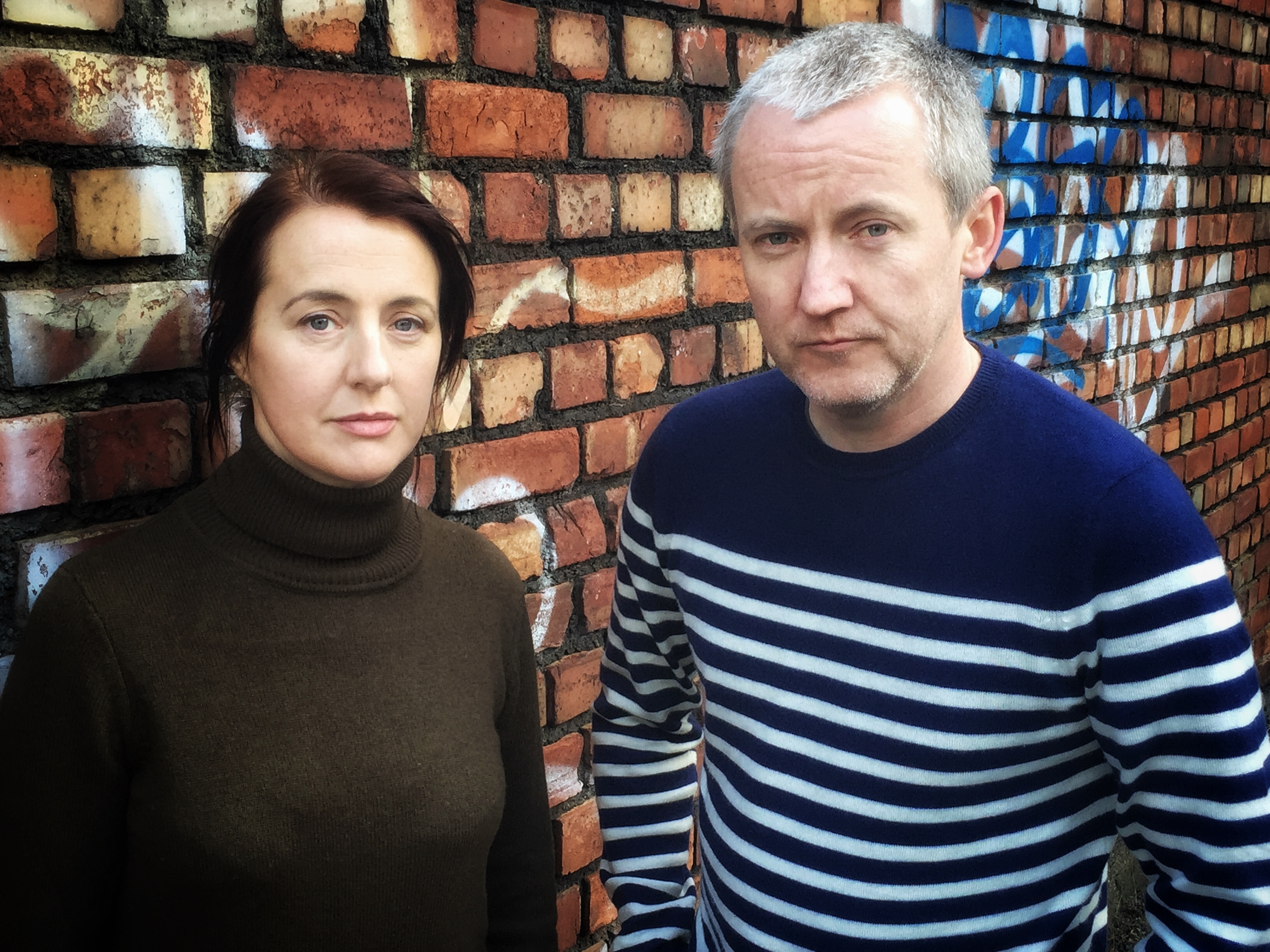 A man and a woman standing side by side against a brick wall, neither smiling but both look content