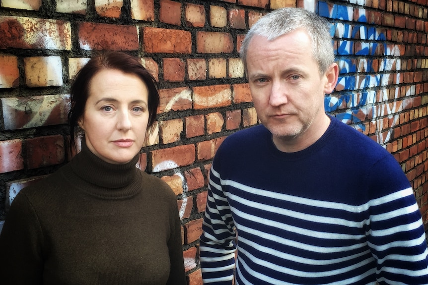 A man and a woman standing side by side against a brick wall, neither smiling but both look content