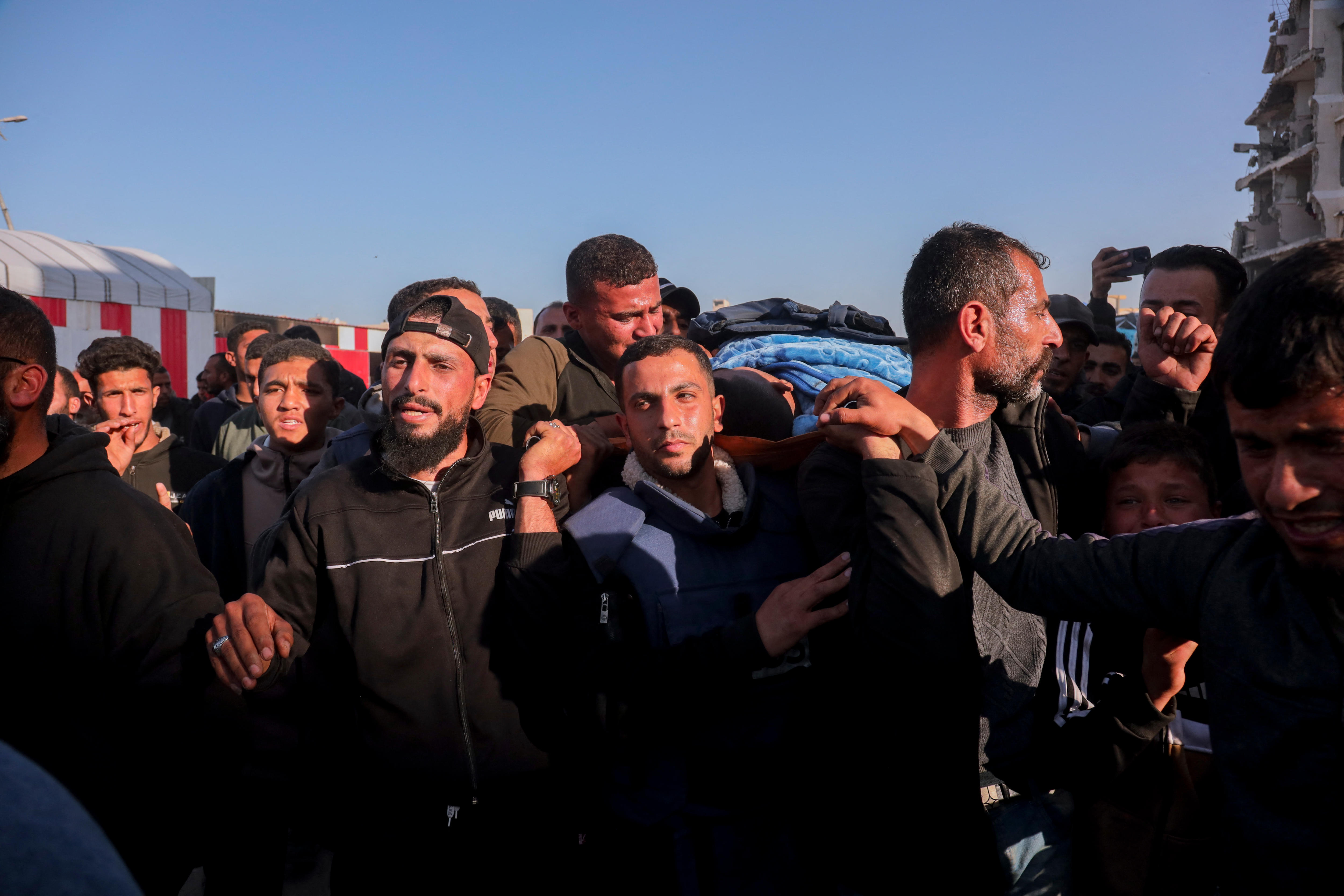 A group of young and middle-aged Palestinian men carry a body on a funeral stretcher through a crowd on a street.