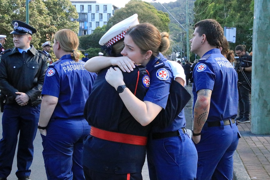 Two paramedics hug wearing their uniforms on a road outside.