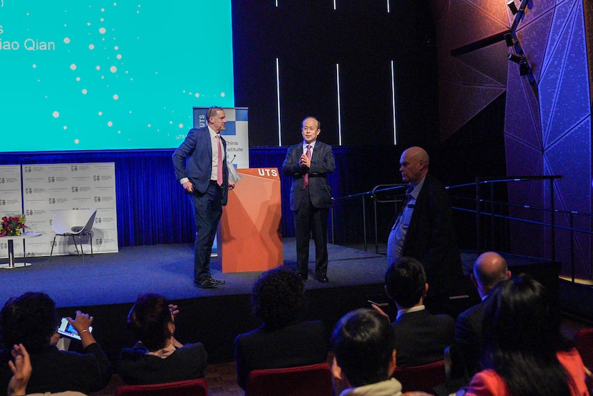 A far view of a stage with the ambassador wearing a suit and a podium reading 'UTS'