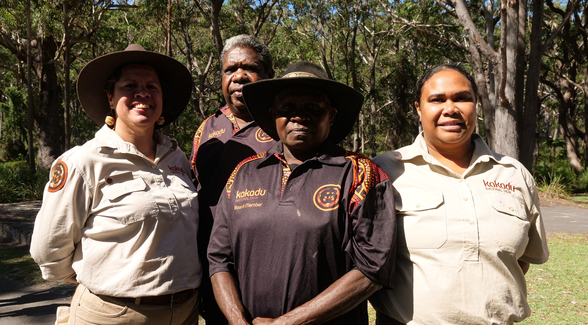 Четыре человека в рубашках цвета хаки с логотипом, двое в коричневых с логотипом и один в шляпе стоят перед деревьями и улыбаются.