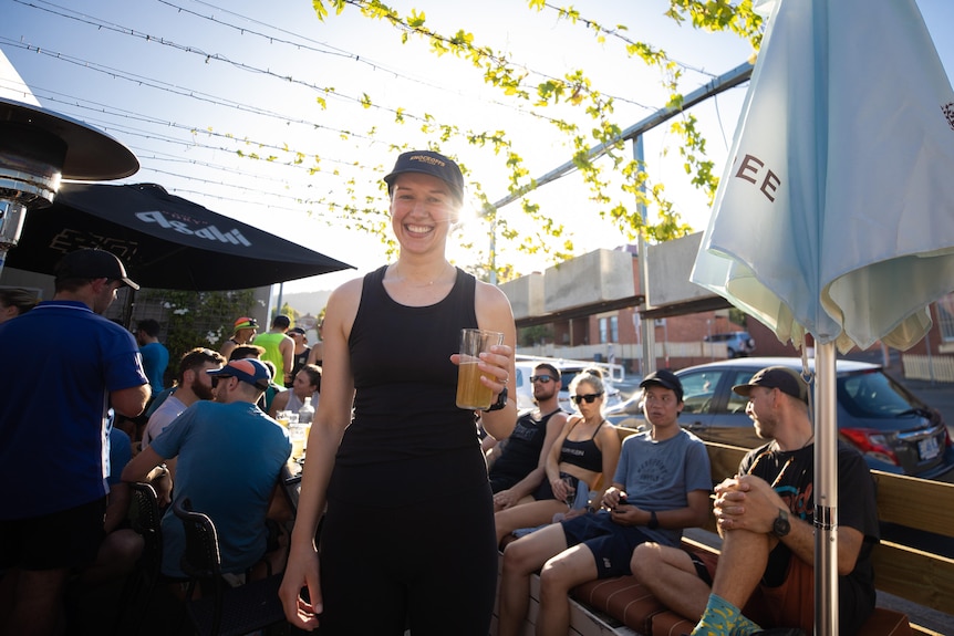 A woman smiles while holding a beer as people sit and stand in the background. 
