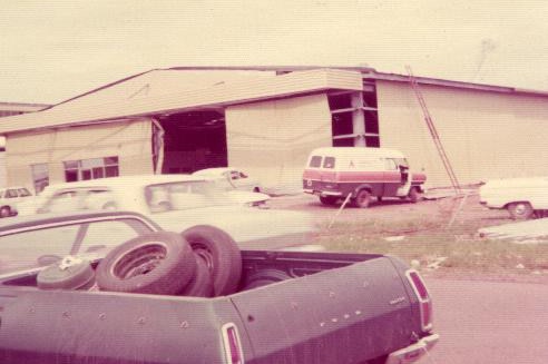An old photo image of a battered aeroplane freight shed with a ute in the frame.