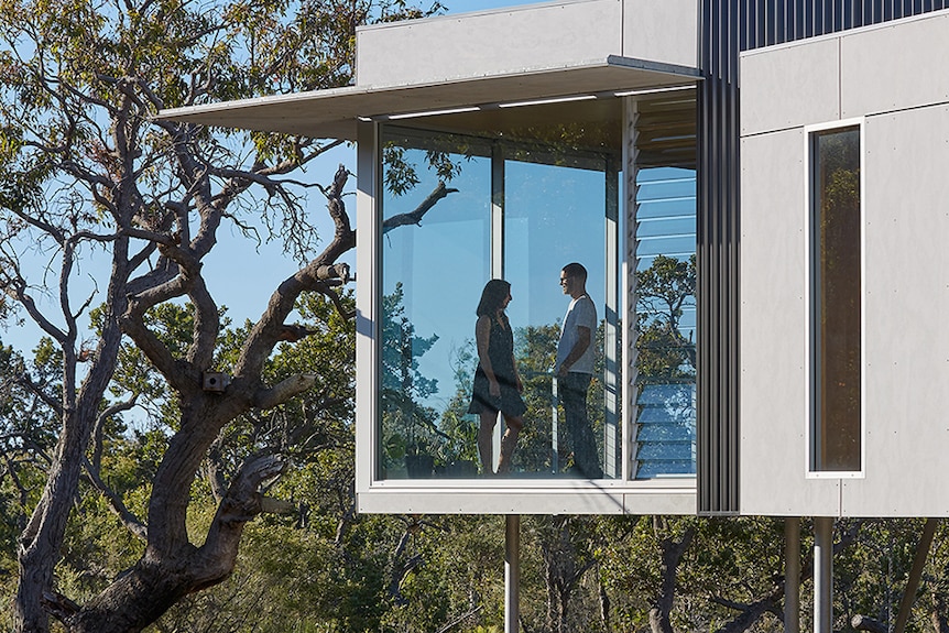 A man and a woman stand behind floor to ceiling windows of a house in the bush. 