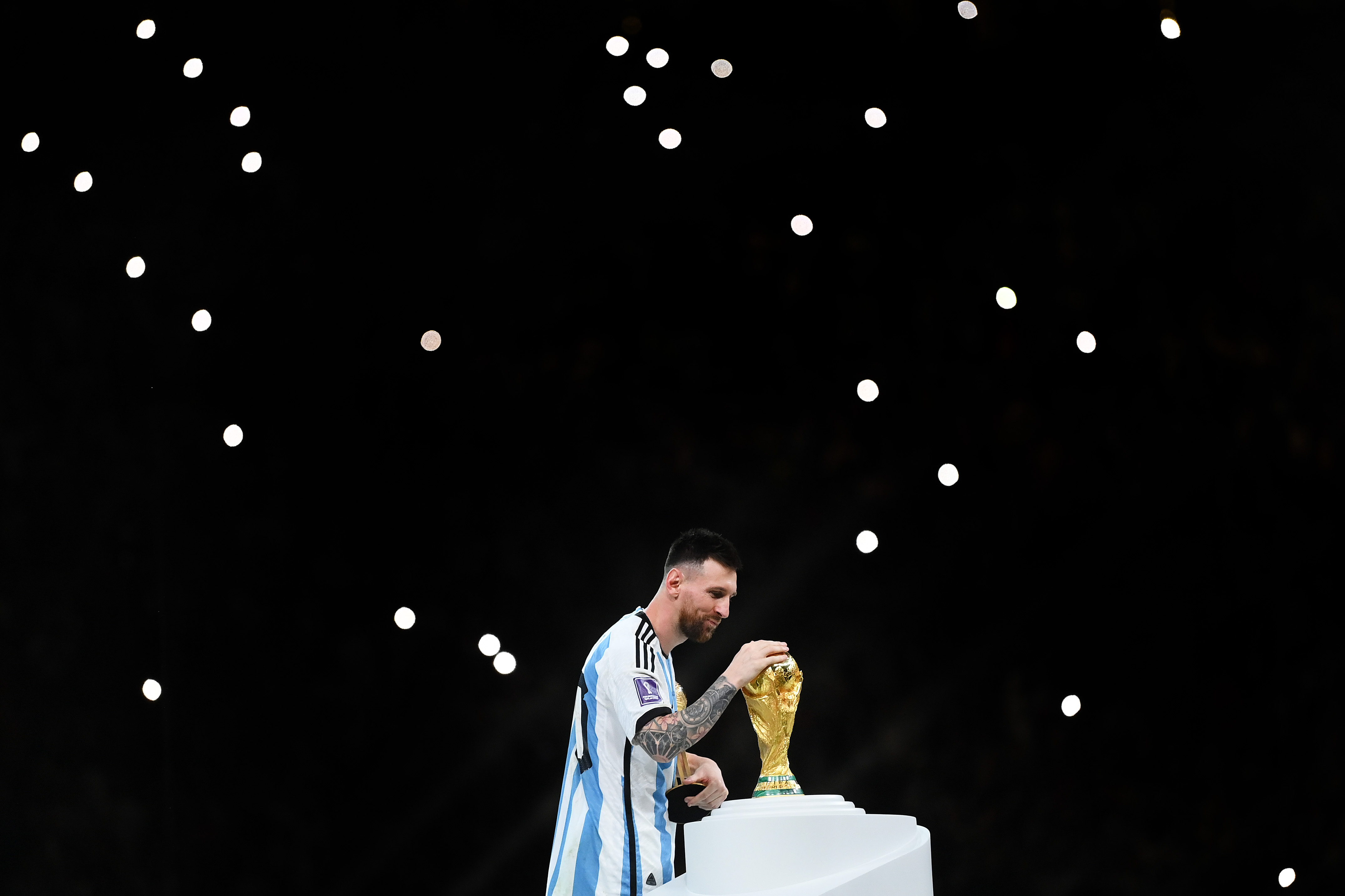 阿根廷的莱昂内尔·梅西 (Lionel Messi) 在赢得卡塔尔决赛后触摸世界杯奖杯，足球能否从自身中拯救出来，背景是摄像机灯。