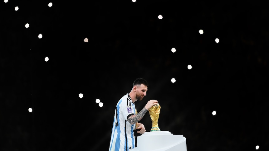 Argentina's Lionel Messi touches the World Cup trophy after winning the Qatar final, with camera lights in the background.