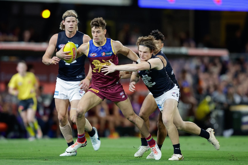 A Brisbane Lions AFL player holds the ball in one hand while trying to fend off a Carlton player with the other.