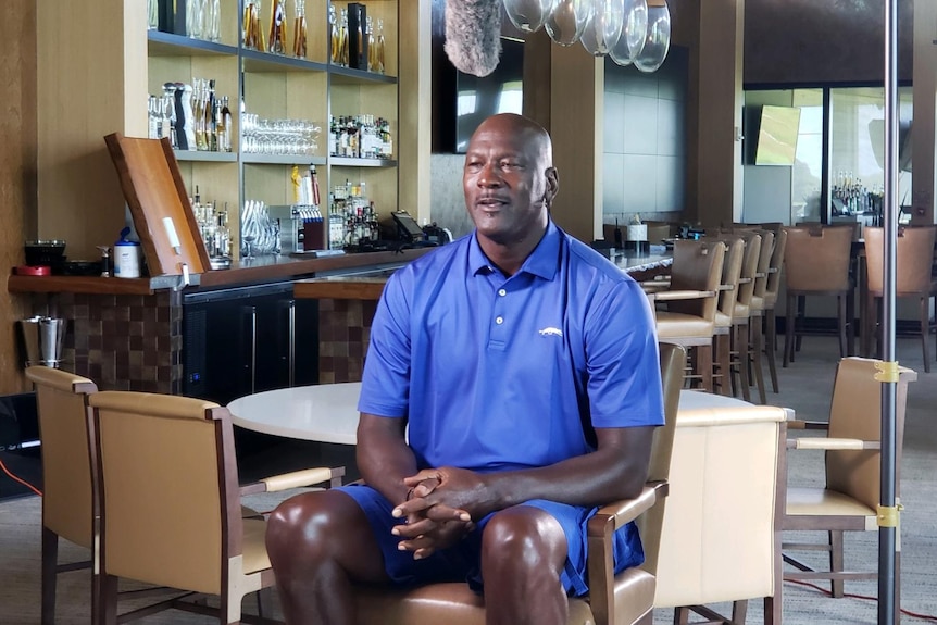 Wide shot Jordan wearing all blue sitting in a chair with golf dining room behind him and TV lights set up around him.