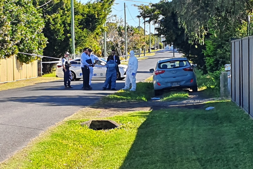 Police and crime scene officers in a street, with a grass nature strip and wooden fence on the right.