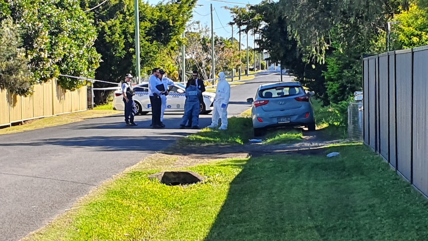 Police and crime scene officers in a street, with a grass nature strip and wooden fence on the right.