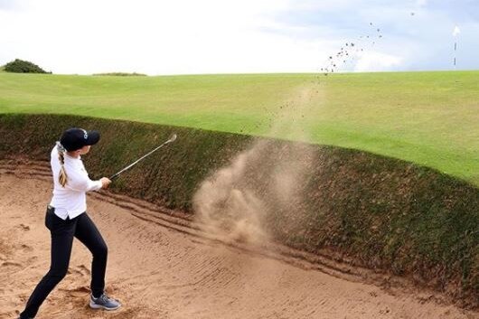Sand sprays in front of a golfer, after she splashes the ball out of a bunker.