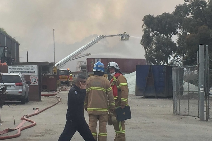 Two firefighters speak outside an industrial site as smoke billows in the background.