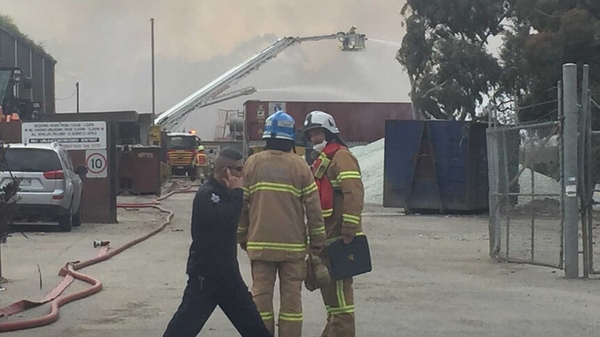Two firefighters speak outside an industrial site as smoke billows in the background.