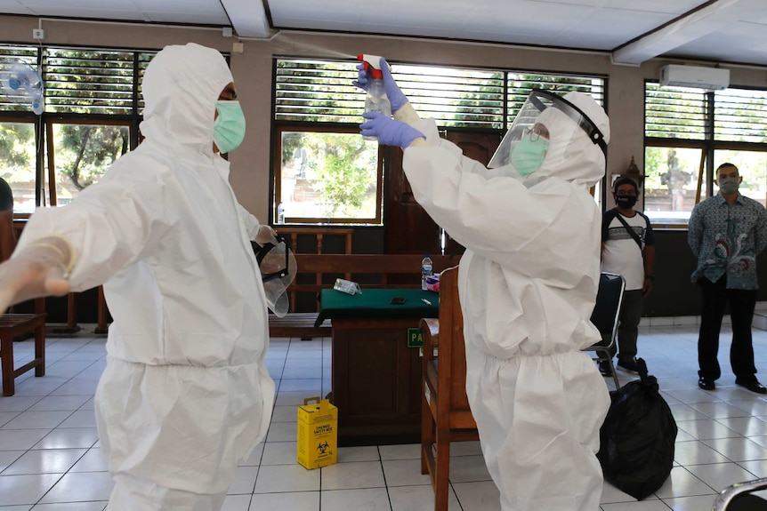 A health worker in protective gear uses a spray bottle to disinfect another health worker who is also wearing protective gear.