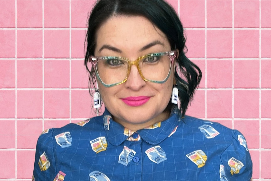 A white, dark-haired woman with large glasses and pink lipstick wearing milk bottle earrings and a patterned short-sleeved shirt