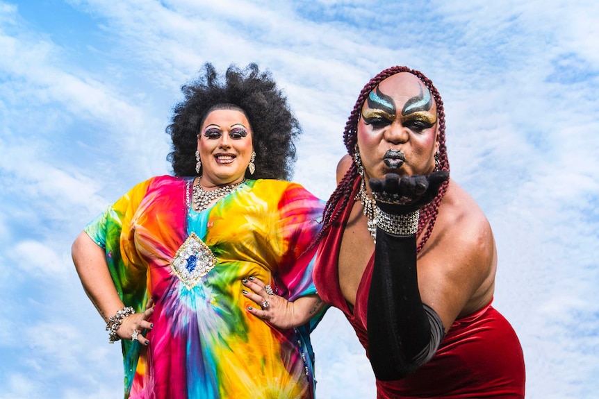 Colour photograph of Lasey Dunaman and Nova Gina of drag act Dreamtime Divas posing with a blue cloudy sky behind them.