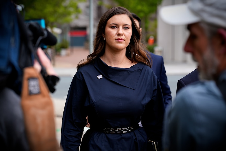 Brittany looks into the camera lens, wearing a navy dress, her hair slightly wavy. Camera operators are on either side of her.