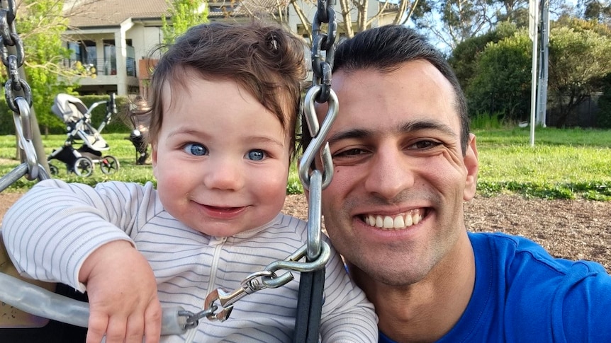Isaac Nowroozi smiles while holding his son Ezra, who is playing on a swing in a park.