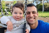 Isaac Nowroozi smiles while holding his son Ezra, who is playing on a swing in a park.