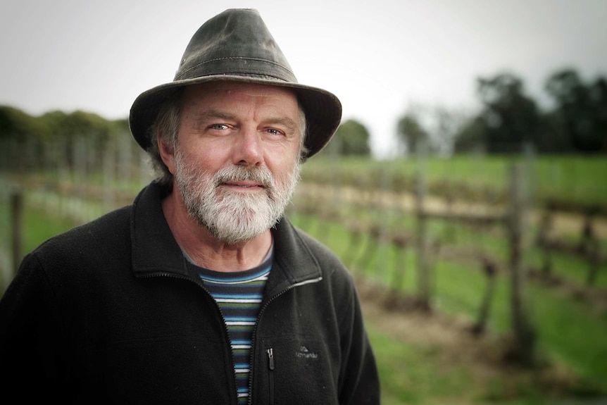 Evan Rolley, a man with a grey-and-black beard and a hat, looks at the camera in front of a vineyard.