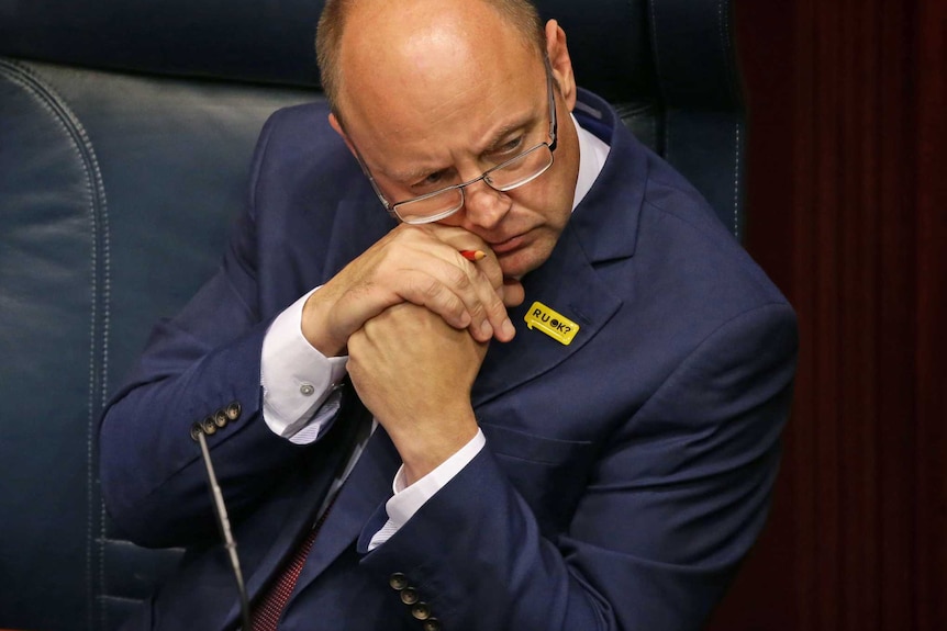 A man in a dark blue suit sits on a navy leather bench in Parliament.